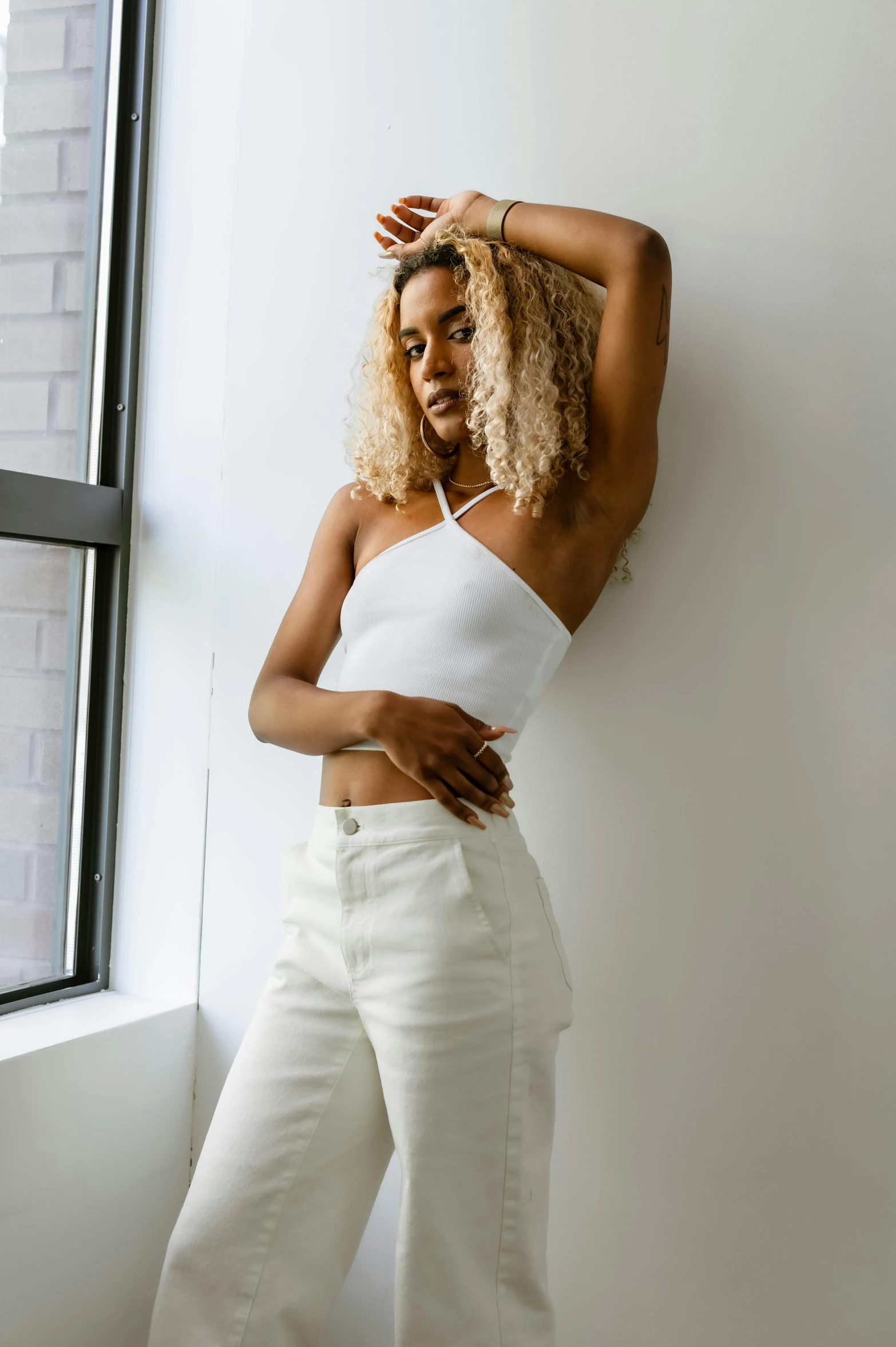 a woman leaning against a wall next to a window, inspired by Esaias Boursse, trending on unsplash, white halter top, white pants, at a fashion shoot, ashteroth