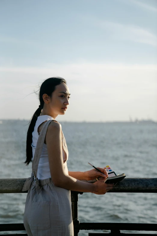 a woman standing in front of a body of water, inspired by Ruth Orkin, pexels contest winner, holding notebook, asian female, looking towards the horizon, near a jetty