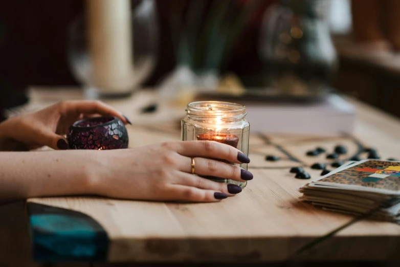 a woman holding a candle on top of a wooden table, by Julia Pishtar, trending on pexels, witchy, with sparkling gems on top, glowing jar, on a white table