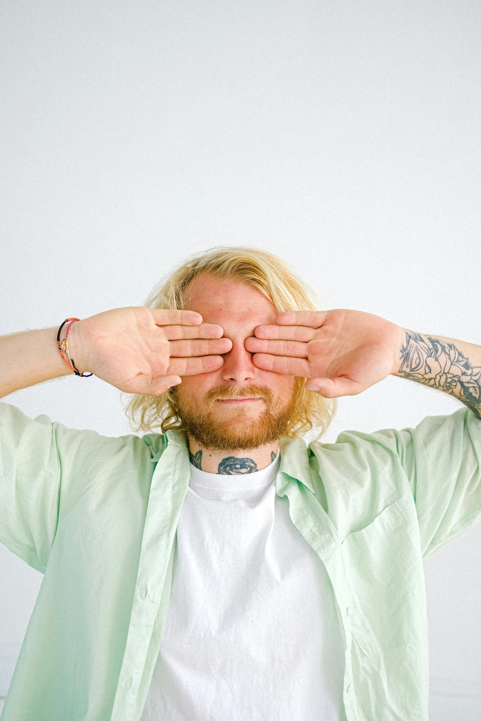 a man covering his eyes with his hands, an album cover, by Jesse Richards, small blond goatee, clear green eyes, tattoos, wearing a light shirt