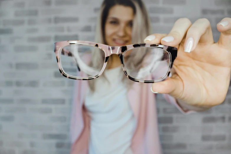 a woman holding up a pair of glasses, a picture, trending on pexels, square glasses, pink glasses, slightly pixelated, professional picture