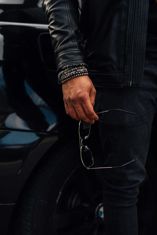 a man in a leather jacket standing next to a car, trending on unsplash, wearing two silver bracelets, wearing black frame glasses, black on black. intricate, profile image