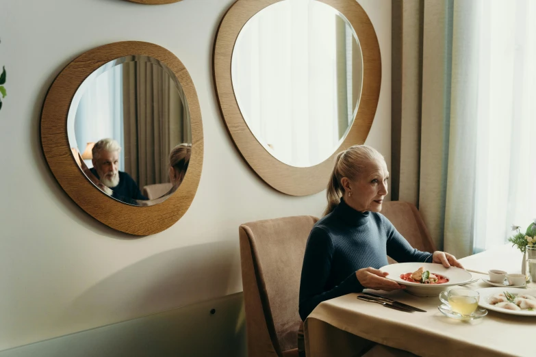 a woman sitting at a table with a plate of food, round mirror on the wall, pierre pellegrini and ash thorp, an elderly, fine dining