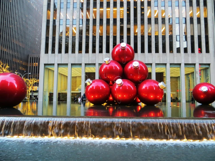 a group of red christmas balls sitting on top of a fountain, inspired by Jeff Koons, new york buildings, mies van der rohe, a cozy, seen from the side