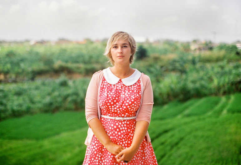 a woman standing on top of a lush green field, an album cover, by Pamela Ascherson, naive art, 50 mm lens photo portrait, wearing red dress, patterned clothing, portrait of kate winslet