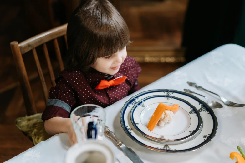 a little girl sitting at a table with a plate of food, by Emma Andijewska, pexels contest winner, arts and crafts movement, gentlemens dinner, white and orange breastplate, place setting, thumbnail