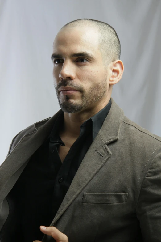 a man in a brown jacket and black shirt, inspired by Carlos Berlanga, dark backdrop, shaven stubble, joseph moncada, on grey background