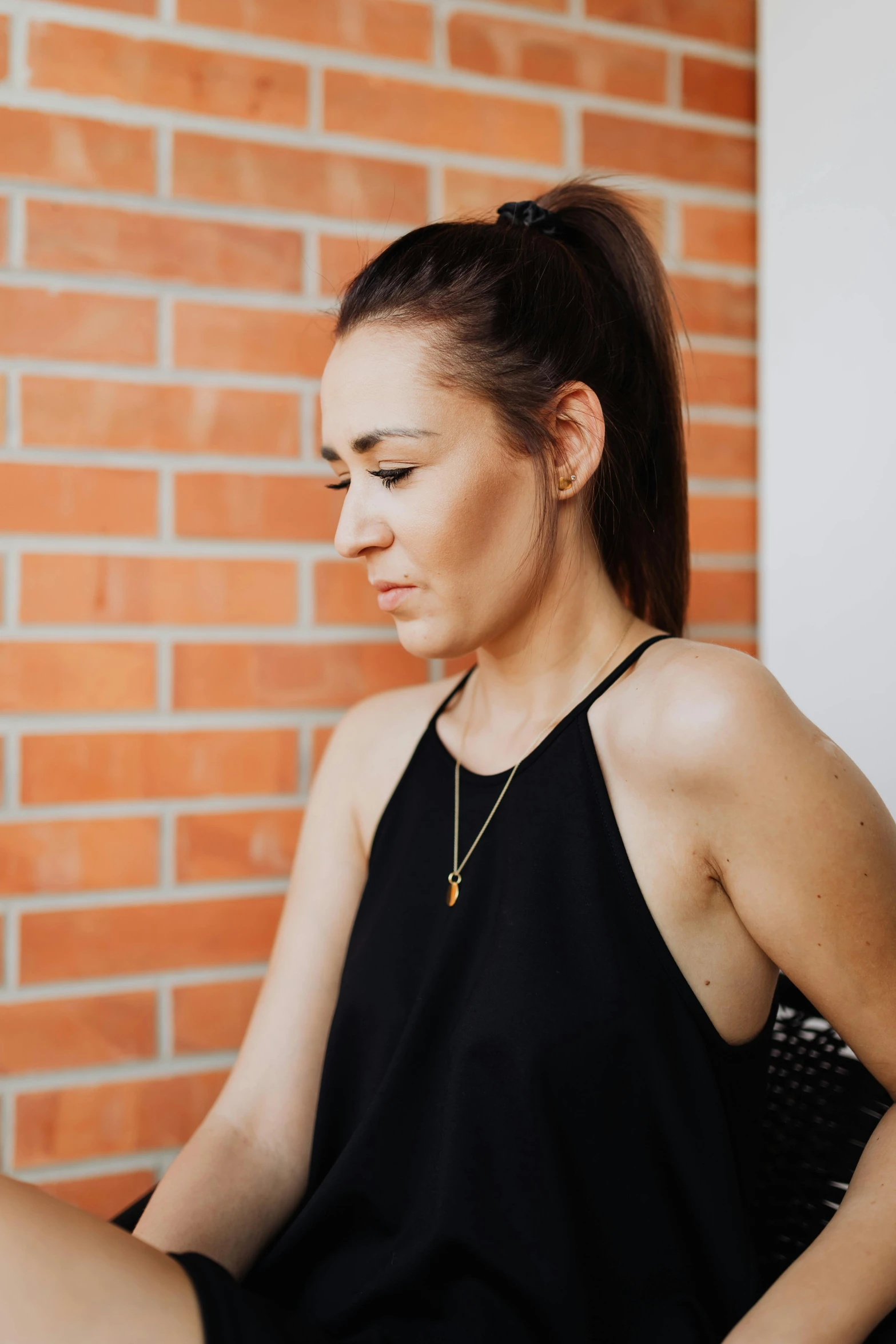 a woman sitting in front of a brick wall, black halter top, profile image, wearing gold jewellery, wearing : tanktop