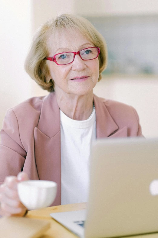 a woman sitting at a table in front of a laptop computer, the look of an elderly person, with a white mug, woman with rose tinted glasses, mobie still