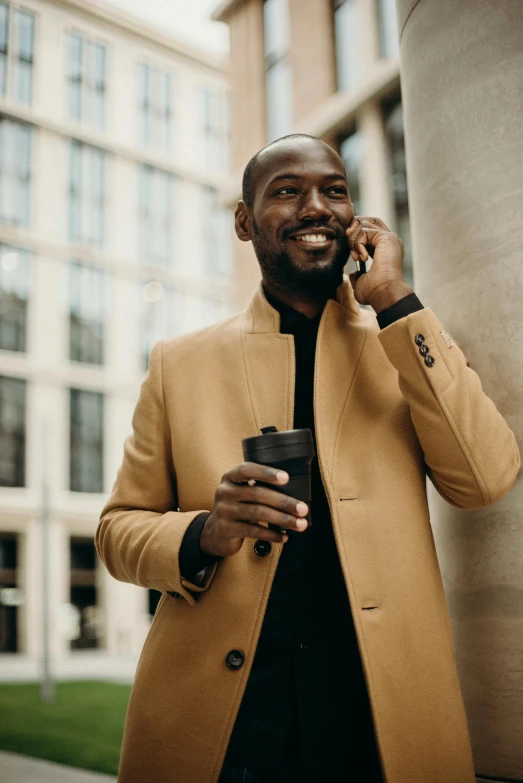 a man standing next to a tall building talking on a cell phone, light brown coat, kevin garnett, high quality product image”, attractive man drinking coffee