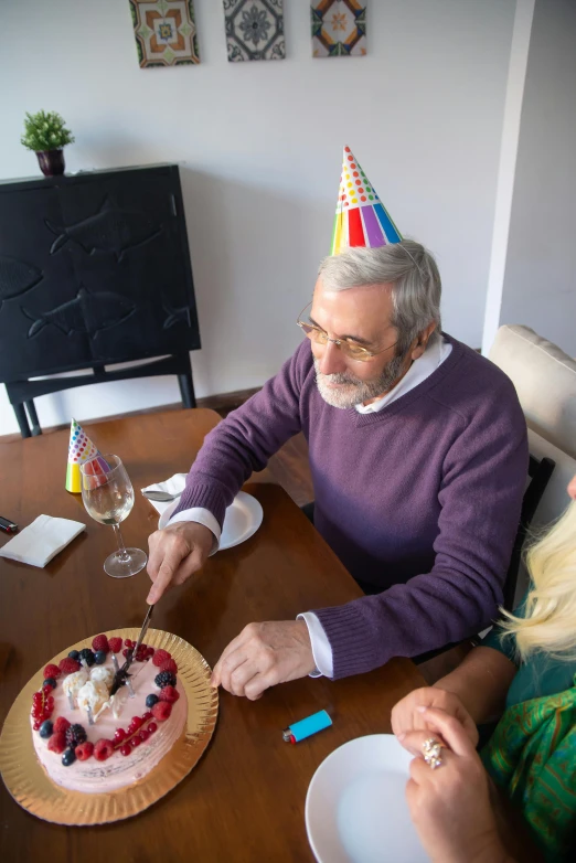 a man and woman sitting at a table with a birthday cake, gray haired, wearing a party hat, 2019 trending photo, apartment