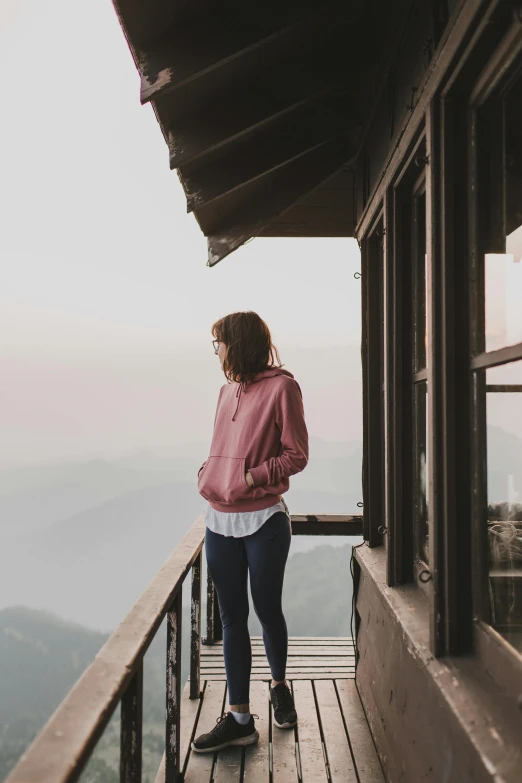 a woman standing on a balcony looking out at the mountains, trending on unsplash, wearing a pink hoodie, full body full height, distant thoughtful look, focused photo