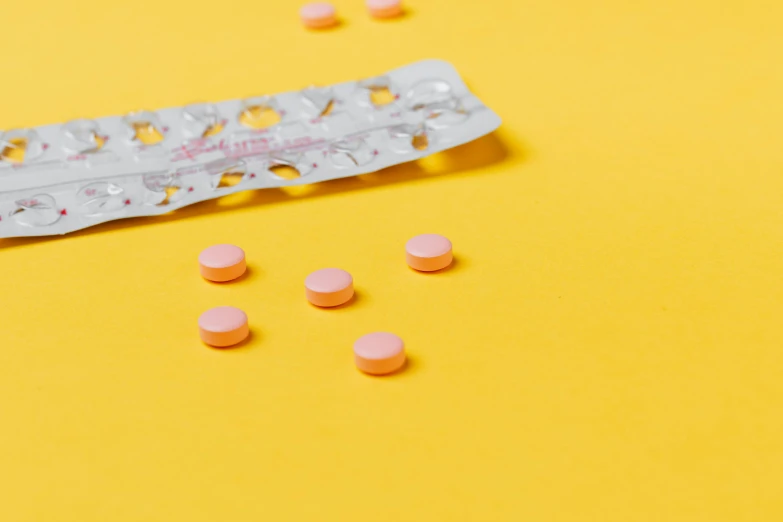 a pack of pink pills sitting on top of a yellow surface, a picture, by Nicolette Macnamara, trending on pexels, contracept, orange pastel colors, shkkeled in the voied, on a table