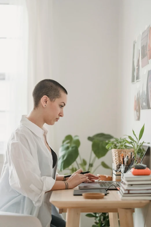 a woman sitting at a table working on a laptop, by Nicolette Macnamara, shaved head, julia sarda, profile image, dwell