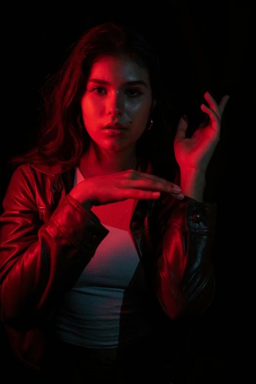 a woman standing in front of a red light, inspired by Elsa Bleda, hand gesture, dark backdrop, red leather jacket, teenage girl