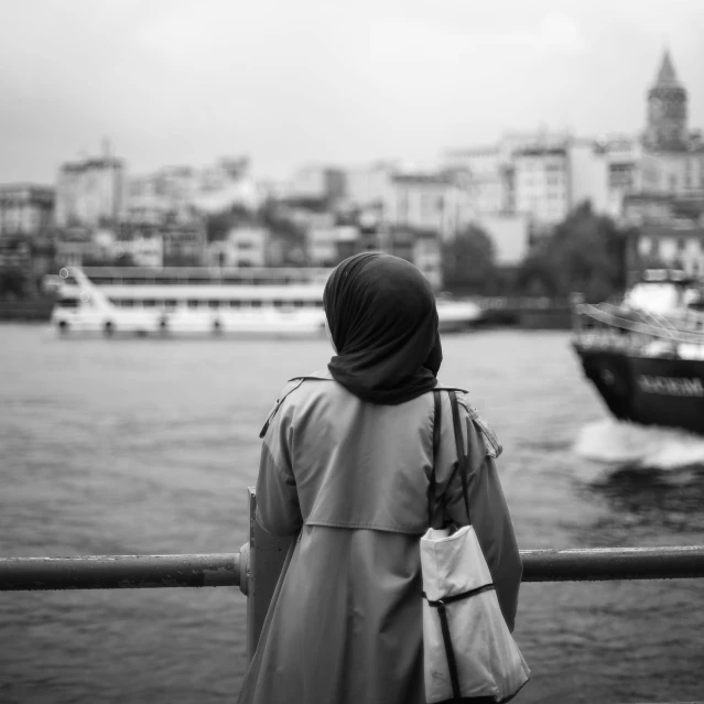 a woman standing in front of a body of water, a black and white photo, by Niyazi Selimoglu, pexels contest winner, hurufiyya, hijab, city views, busy but lonely, 15081959 21121991 01012000 4k