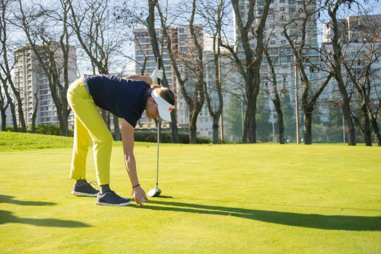 a woman standing on top of a green field holding a golf club, in a city park, profile image, bent over posture, jakub rebelka