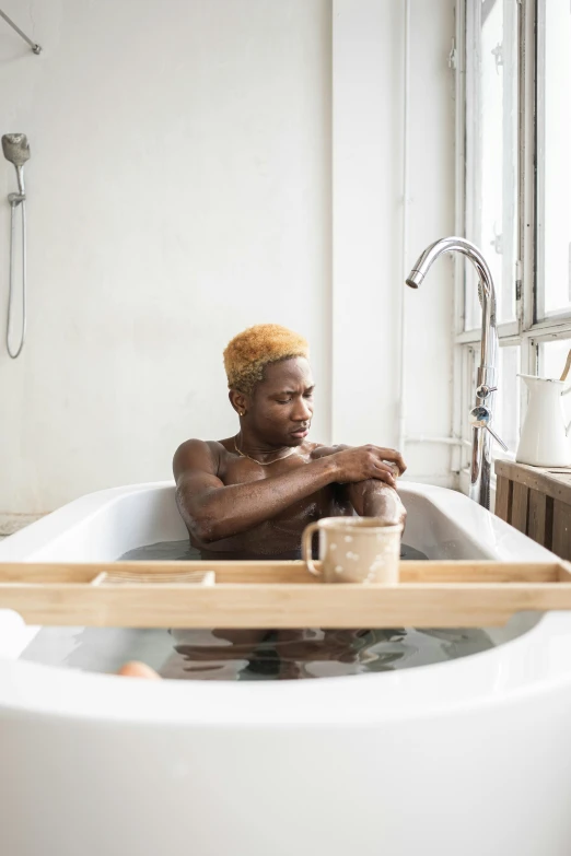 a man sitting in a bathtub next to a window, by Jessie Alexandra Dick, trending on unsplash, short blonde afro, swimming in a pool of coffee, transgender, photographed for reuters