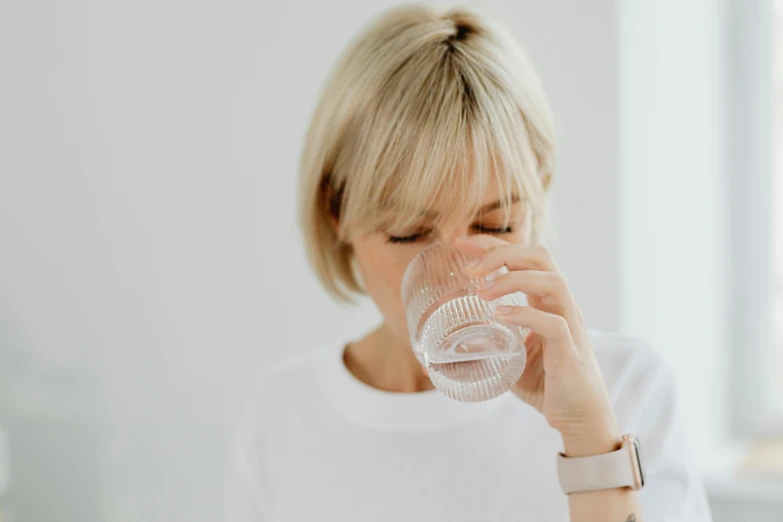 a woman drinking a glass of water, by Nicolette Macnamara, pexels contest winner, a blond, plain background, head to waist, slightly pixelated