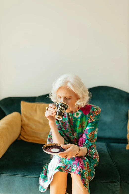 a woman sitting on a couch holding a camera, secret tea society, silver haired, green and brown clothes, woman drinking coffee