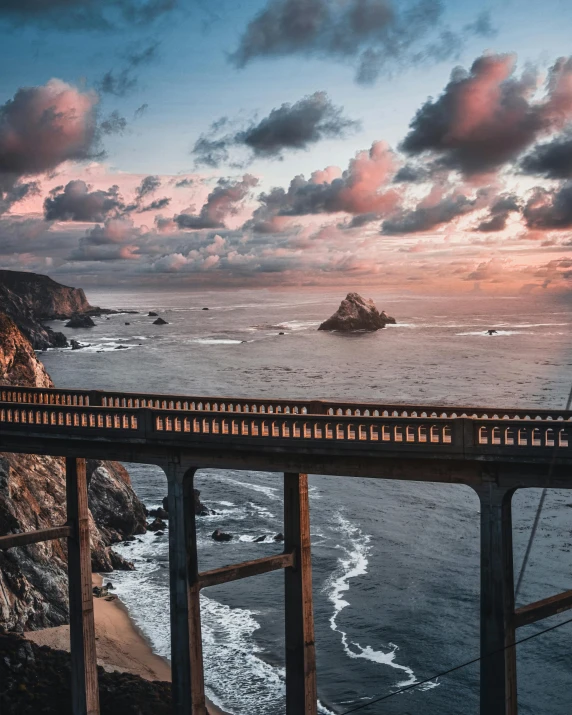 a bridge over a body of water under a cloudy sky, a matte painting, unsplash contest winner, california coast, pink golden hour, steep cliffs, conde nast traveler photo