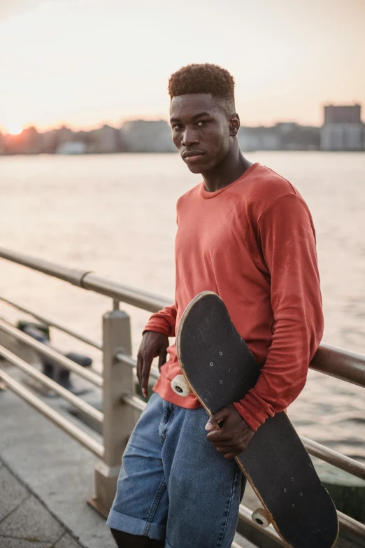 a man standing next to a body of water holding a skateboard, brown skinned, red sweater and gray pants, brooklyn, portrait featured on unsplash