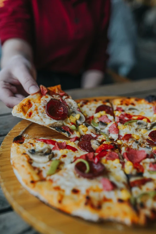 a pizza sitting on top of a wooden cutting board, hand on table, offering a plate of food, multicoloured, woodfired