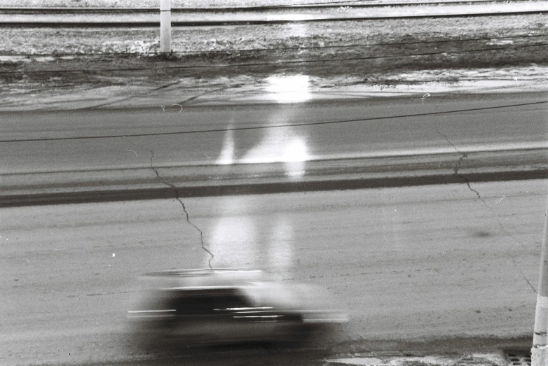 a black and white photo of a car driving down a street, inspired by Louis Faurer, photorealism, wet highway chase, nascar race, 1987 photograph, detail on scene