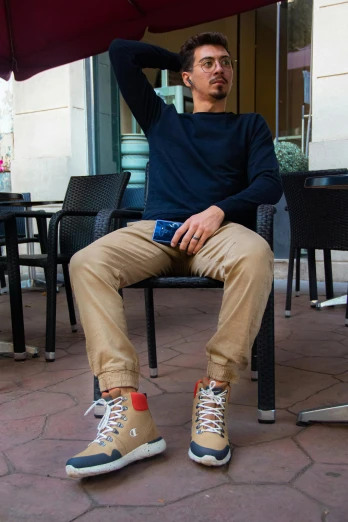 a man sitting on a chair under an umbrella, wearing red converse shoes, khakis, in a navy blue sweater, david luong