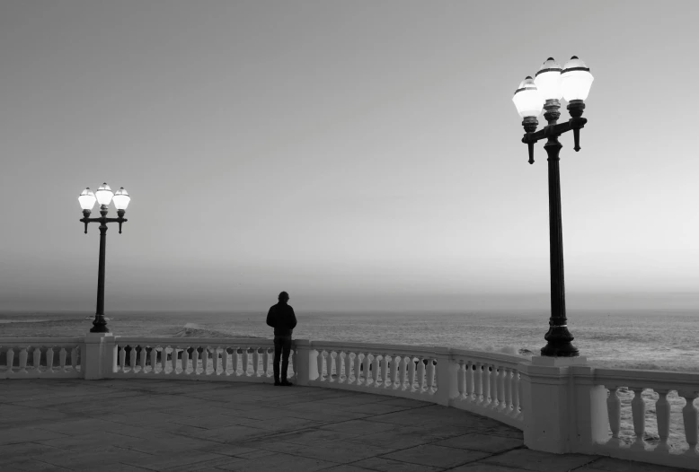 a black and white photo of a person looking out at the ocean, by Giorgio Cavallon, pexels contest winner, romanticism, big beautiful street lamps, at the terrace, calm evening, man