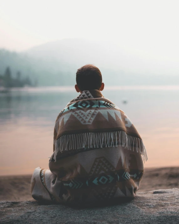 a man sitting on top of a rock next to a body of water, covered with blanket, lgbtq, facing away, instagram post