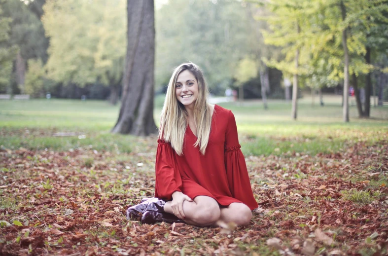 a woman in a red dress sitting on the ground, by Alice Mason, pexels contest winner, antipodeans, smiling young woman, autum, at a park, italian looking emma