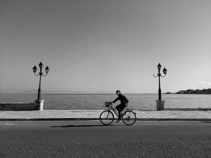 a man riding a bike down a street next to the ocean, a black and white photo, by Alexis Grimou, pexels contest winner, minimalism, greece, ffffound, venice, various posed