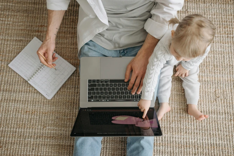 a man sitting on the floor with a baby using a laptop, a cartoon, pexels, digital oth, top - down photo, middle of the page, 1 2 9 7