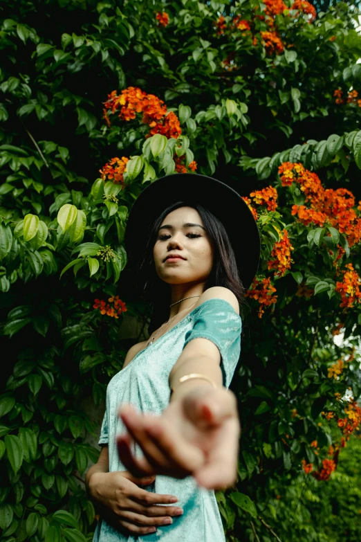 a woman in a hat standing in front of a tree, an album cover, by Robbie Trevino, pexels contest winner, picking up a flower, orange and teal color, beautiful young asian woman, pov photo