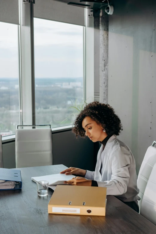 a woman sitting at a table reading a book, by Lily Delissa Joseph, pexels contest winner, modernism, set inside of office, corporate memphis, nathalie emmanuel, sitting on top a table