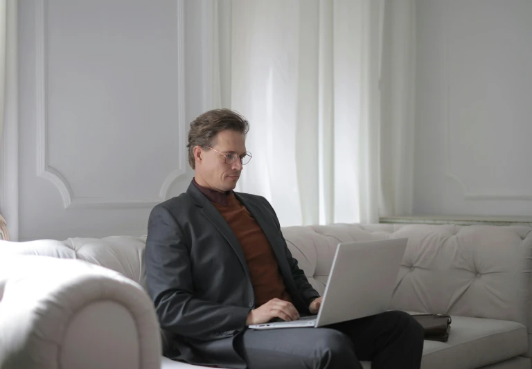 a man sitting on a couch using a laptop computer, pexels, formal attire, paul barson, in a white room, lachlan bailey