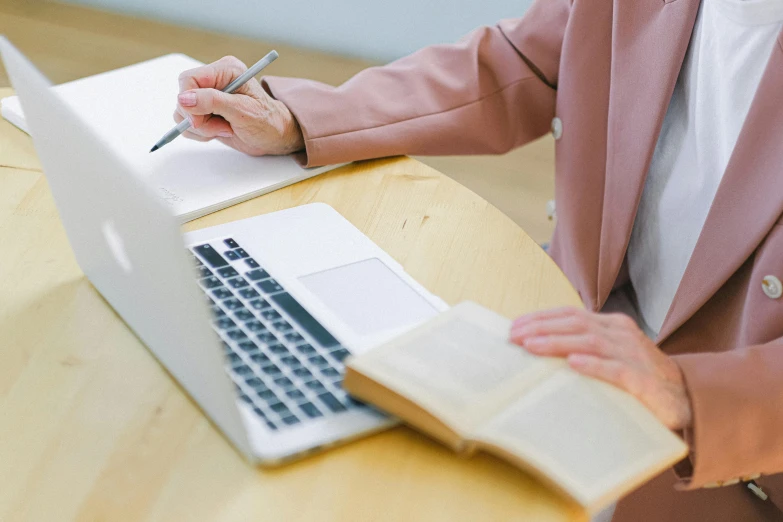 a woman sitting at a table with a laptop and a notebook, by Nicolette Macnamara, trending on pexels, librarian, holding pencil, clean thick line, brown