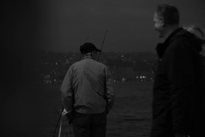 a couple of men standing next to each other near a body of water, a black and white photo, by Tamas Galambos, night time footage, fishing, istanbul, [ cinematic