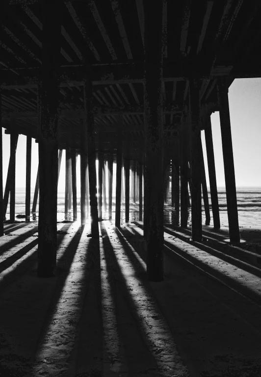 a black and white photo of the underside of a pier, deep shadows and colors, square lines, sunshafts, california;