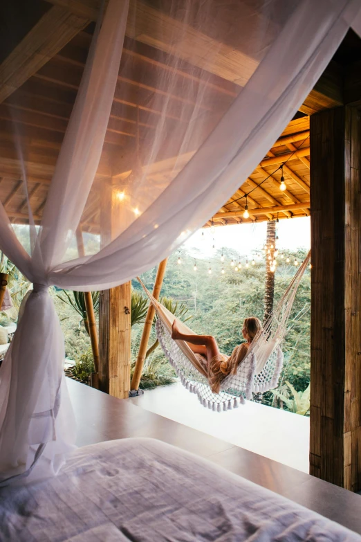 a woman laying in a hammock on top of a bed, by Jessie Algie, unsplash contest winner, light and space, floral jungle treehouse, luxurious wooden cottage, panoramic shot, high view