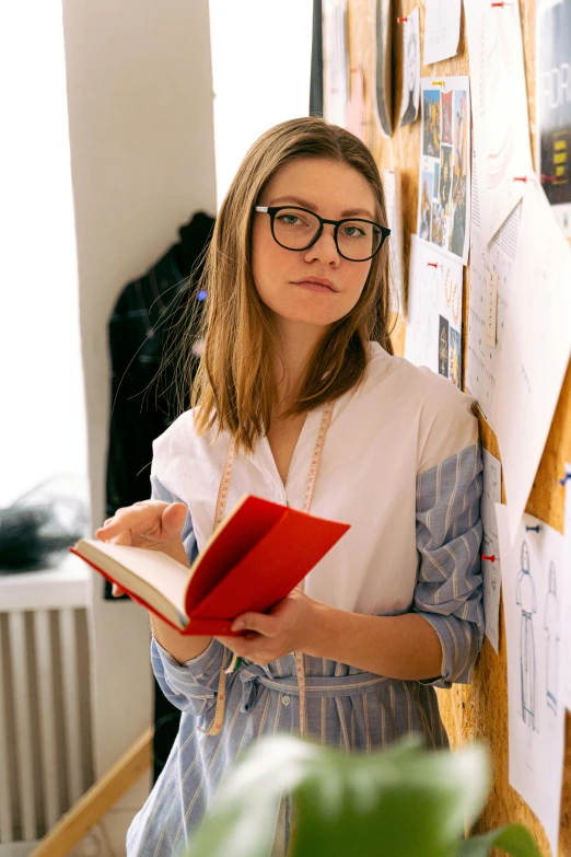 a woman standing in front of a bulletin board holding a book, a picture, by karolis strautniekas, trending on unsplash, academic art, wearing square glasses, professional model, looking serious, fashion designer