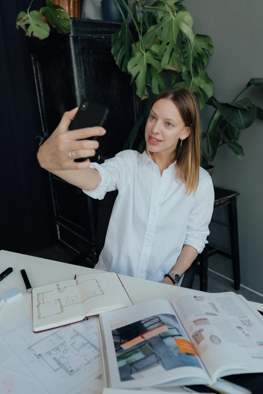 a woman sitting at a table taking a selfie, architectural planning, style of julia razumova, standing on a desk, holding it out to the camera