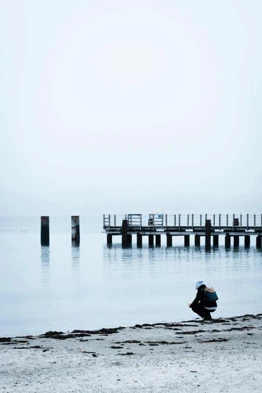 a person sitting on a beach next to a body of water, inspired by Pierre Pellegrini, unsplash, minimalism, pale blue fog, docks, crouching, winter photograph