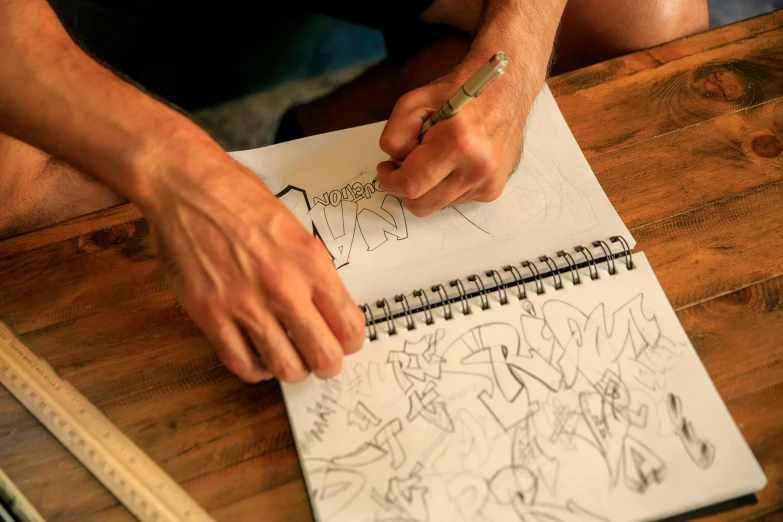 a man sitting at a table drawing on a piece of paper, a drawing, by Lee Loughridge, pexels, process art, graffiti letters, holding notebook, dale keown and van sciver, album cover