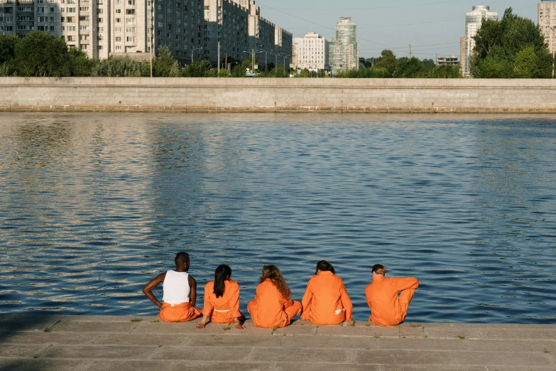 a group of people sitting next to a body of water, by Attila Meszlenyi, wearing orange prison jumpsuit, moscow, city morning, young girls