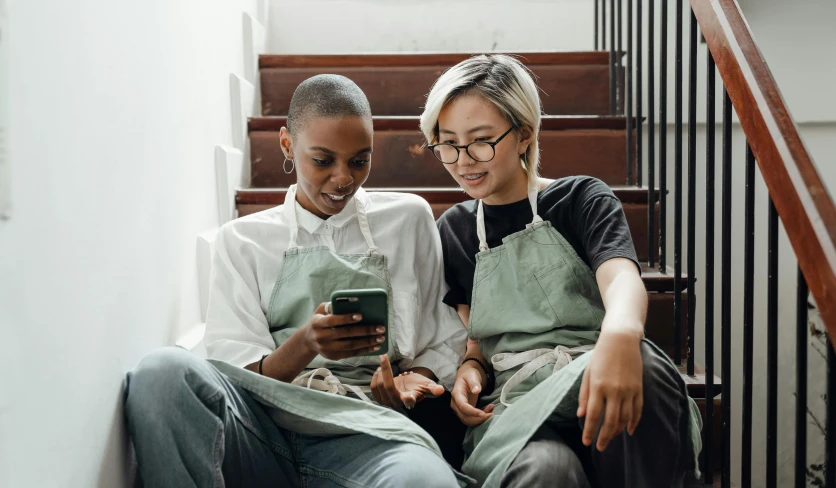 two women sitting on stairs looking at a cell phone, by Emma Andijewska, trending on pexels, happening, wearing an apron, aussie baristas, mix of ethnicities and genders, embracing