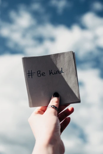 a person holding a piece of paper with the word be kind written on it, by Niko Henrichon, trending on unsplash, skies, 256435456k film, ribbon, grey