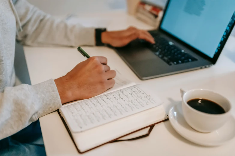 a person sitting at a desk with a laptop and a cup of coffee, by Carey Morris, trending on pexels, ancient writing, holding notebook, background image, caligraphy