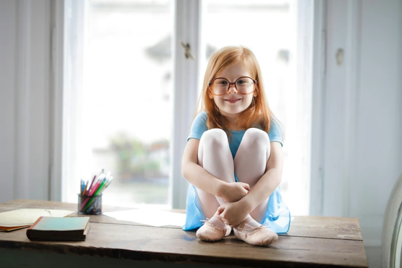 a little girl sitting on top of a wooden table, pexels, square rimmed glasses, she is smiling, ballerina, glasses |